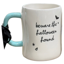 Load image into Gallery viewer, A white ceramic mug with a light blue handle, featuring the black cursive text 'beware the halloween hound' along with candy corn and star illustrations. A black ribbon is tied to the handle, and the mug is photographed on a plain white background.
