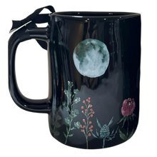 Load image into Gallery viewer, A glossy black ceramic mug featuring a printed design of a large pale moon and botanical illustrations, including a pink flower and green leaves, with a black ribbon tied to the handle.
