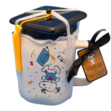 Load image into Gallery viewer, White mug that shows snoopy jumping upside down with a blue graduation gown on with a black lid that is shaped like a graduation cap.
