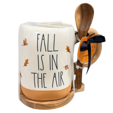 Load image into Gallery viewer, A ceramic mug with a wooden spoon and wooden stand, featuring the text 'FALL IS IN THE AIR' and small autumn leaves with a tan handle and bottom on a white background, with a hanging orange tag. 

