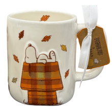 Load image into Gallery viewer, A white ceramic Snoopy mug featuring Snoopy lying on an orange and brown plaid doghouse with scattered autumn leaves. A white ribbon and a brown Peanuts hangtag are attached to the handle.

