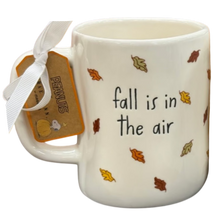 Load image into Gallery viewer, A white ceramic mug with the phrase 'fall is in the air' printed in black, decorated with small autumn leaves in orange, yellow, and brown. A white ribbon and a brown tag tied to the handle read 'PEANUTS' 
