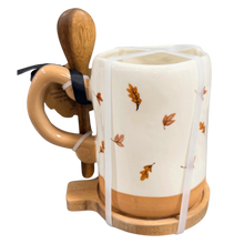 Load image into Gallery viewer, A ceramic mug with a cream-colored body, tan bottom and handle, decorated with pressed autumn leaves, a light wood handle, a wooden spoon, and a white protective wrapping strap.
