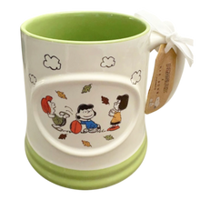 Load image into Gallery viewer, A ceramic mug featuring the female Peanuts characters playing with a football, with a light green interior and base, a white exterior, and a wooden hang tag.
