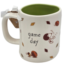 Load image into Gallery viewer, A white ceramic coffee mug with a green interior, decorated with Snoopy illustrations, autumn leaves, and the words 'game day' in black script. A white ribbon and small tag are tied to the handle.
