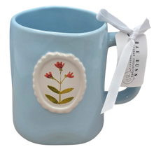 Load image into Gallery viewer, Light blue mug with circular floral design and ribbon tag on a white background
