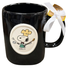 Load image into Gallery viewer, Black mug with Snoopy cartoon character design dressed as a chef and a white ribbon on a white background circle on a black mug.
