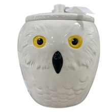 Load image into Gallery viewer, A white ceramic owl-shaped jar with a matching lid, featuring yellow eyes, a black beak, and a small white ribbon tag attached at the top.
