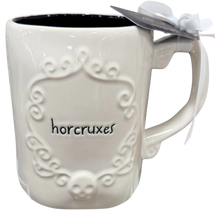 Load image into Gallery viewer, An off-white ceramic mug with a black interior, featuring an embossed gothic-style crest with a skull at the bottom and the word 'horcruxes' in black script. A white ribbon is tied to the handle, and a small silver tag is attached.

