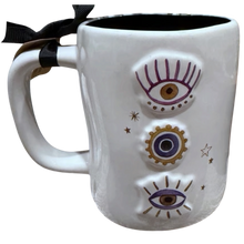 Load image into Gallery viewer, A white ceramic mug with black interior, decorated with three vertical evil eye illustrations in purple, gold, and blue, with small gold stars.
