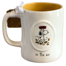 Load image into Gallery viewer, A white ceramic coffee mug with a yellow interior, featuring a Snoopy illustration in the air with falling leaves and the text 'in the air' printed below. A brown gift tag with a white ribbon is attached to the handle.
