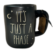 Load image into Gallery viewer, A black ceramic mug with the phrase 'IT'S JUST A PHASE' in cream-colored lettering, featuring a small moon and star graphic. A brown paper hangtag is attached to the handle.
