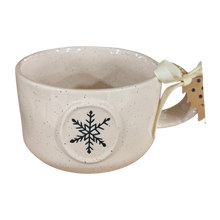 Load image into Gallery viewer, An off-white speckled ceramic mug with a black snowflake design on the front and a cream ribbon bow with a paper tag attached to the handle, shown on a plain white background.

