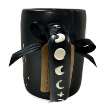 Load image into Gallery viewer, A black ceramic mug with a sculpted handle featuring moon and star motifs, tied with a black ribbon and a brown paper tag, on a white background.
