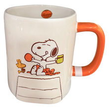 Load image into Gallery viewer, A white ceramic mug featuring a Snoopy and Woodstock illustration, with a bright orange handle and matching orange details, shown on a plain white background.
