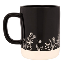 Load image into Gallery viewer, A black ceramic mug with a cream-colored base and delicate white floral line art illustration around the lower half, shown against a plain white background.

