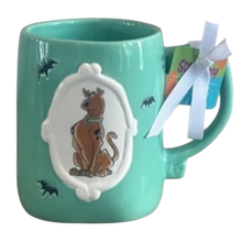 Load image into Gallery viewer, A turquoise ceramic mug featuring a 3D relief of Scooby-Doo inside a white decorative frame, decorated with small black bat silhouettes. A white ribbon is tied to the handle, holding colorful tags.
