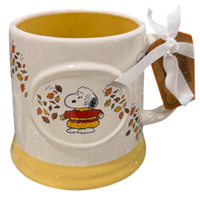 Load image into Gallery viewer, A ceramic mug featuring Snoopy in a red and yellow sweater surrounded by falling autumn leaves, with a yellow interior and base, a white ribbon, and a Peanuts tag on the handle.
