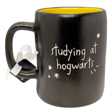 Load image into Gallery viewer, A black ceramic mug with a yellow interior, featuring the text 'studying at hogwarts' in white script with small white star and punctuation marks. A white ribbon is tied around the handle, and the mug is photographed on a clean white background.
