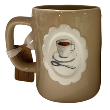 Load image into Gallery viewer, A glossy ceramic coffee mug in a warm taupe-brown color, featuring a white scalloped oval medallion on the front with a painted illustration of a coffee cup, saucer, and spoon. A white ribbon with a brown paper tag is tied to the handle.
