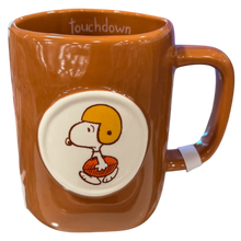 Load image into Gallery viewer, A brown ceramic mug featuring a circular white insert with the Peanuts character Snoopy wearing a yellow football helmet. The word 'touchdown' is printed inside the mug.
